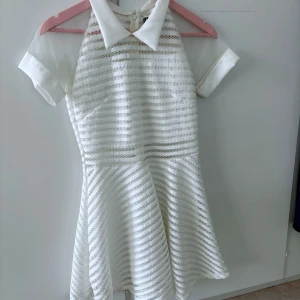 Vit kortklänning med krage från LOX - Säljer en söt vit kortklänning från LOX med klassisk skjortkrage och korta ärmar. Klänningen har ett diskret randigt mönster i tyget och dragkedja bak. Perfekt för dig som gillar en stilren och fräsch look.