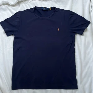 Mörkblå t-shirt från Polo Ralph Lauren - Kostar 1200 ny. Passar 13-14 åringar. Säljer en mörkblå t-shirt från Polo Ralph Lauren med klassisk broderad logga på bröstet. Modellen är custom slim fit och har rund hals och korta ärmar. Perfekt för dig som gillar stilrena basplagg med en sportig touch.