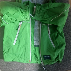 Grön vindjacka från Polarn O. Pyret - Snygg grön vindjacka med huva från Polarn O. Pyret. Jackan har vita dragkedjor framtill och på fickorna samt reflexdetalj på ryggen. Meshfoder på insidan för extra komfort. Perfekt för utomhusaktiviteter.jackan är i storlek 92!