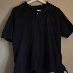 Mörkblå pikétröja från Polo Ralph Lauren - Säljer en klassisk mörkblå pikétröja från Polo Ralph Lauren med liten röd logga broderad på bröstet. Tröjan har krage, knappar vid halsen och är kortärmad. Perfekt för en stilren och avslappnad look.