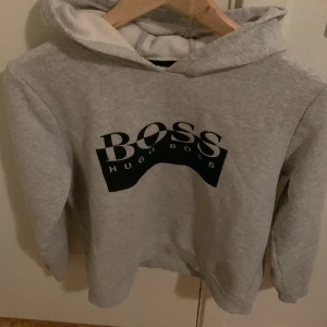 Grå hoodie från Hugo Boss - Säljer en grå hoodie från Hugo Boss med stor svart logga på bröstet. Tröjan har huva och är fodrad med mjukt material på insidan. Perfekt för en avslappnad och stilren look. Du ansvarar för frakt.