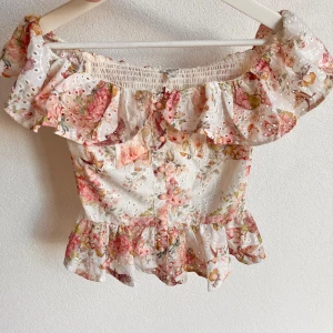 Blommig topp - Superfin blommig offshoulder topp från River Island med volangdetaljer och smockad rygg. Topp i vitt med rosa, gröna och gula blommor, broderade hål och volanger både upptill och nertill. Perfekt för varma dagar!