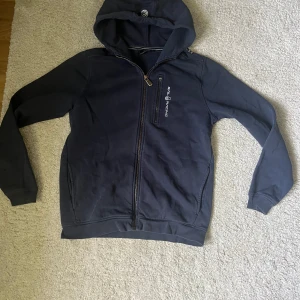 Mörkblå hoodie från Sail Racing - Mörkblå hoodie från Sail Racing med dragkedja framtill och en bröstficka med dragkedja. Tröjan har huva och broderad logga på bröstet. Perfekt för kyliga dagar och enkel att matcha med andra plagg. Kedjan har gått av på lilla fickan. Annars Nytt skick. Barnstorlek 170 motsvarar XS och KANSKE S i vuxen storlek.