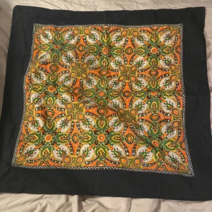 Färgglad mönstrad bandana med svart kant - Säljer en fyrkantig bandana med färgstarkt mönster i orange, grönt och gult samt svart ram runtom. Mönstret är detaljerat och symmetriskt, vilket ger en retrovibe. Perfekt accessoar för att liva upp din outfit.