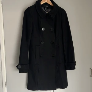 Svart ullkappa från H&M 36 - Stilren svart A-linje skuren ullkappa från H&M storlek 36 med dubbelknäppning och stora knappar framtill. Kappan har klassisk krage, långa ärmar och markerad midja med dekorativ detalj bak. En av fickorna fram har lagats och sytts ihop i fodret och knapparna på midjedetaljen har tagits bort. 