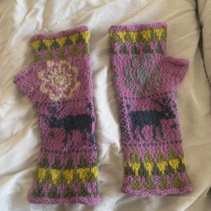 Stickade tumlösa vantar med älgmotiv - Unika stickade vantar i lila, grönt och gult med tumhål och älgmotiv på framsidan. Mönstrade detaljer och blommor ger en färgglad och personlig stil. Perfekta för dig som vill ha något annorlunda och mysigt.