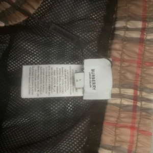 Rutigta badshorts från Burberry - Säljer ett par klassiska badshorts från Burberry i beige med det ikoniska rutmönstret. Shortsen har elastisk midja och innerbyxa i mesh. Perfekta för sommaren och strandhäng.