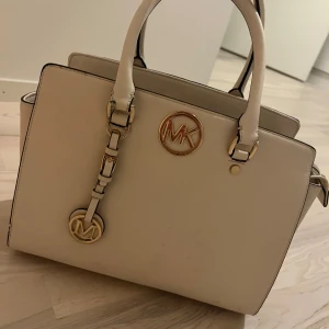 Vit handväska från Michael Kors - Snygg och stilren vit handväska från Michael Kors med guldfärgade detaljer och MK-logga framtill. Väskan har två handtag, dekorativt hänge och innerfack med dragkedja. Perfekt accessoar för att lyfta din outfit.