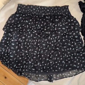 Svart blommig volangkjol - Supersöt svart kjol med små blommor i vitt och rosa. Kjolen har flera lager volanger och resår i midjan för en bekväm passform. Perfekt att styla med en stickad tröja eller en enkel topp.