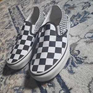 Vans slip-on checkerboard sneakers - Klassiska Vans slip-on sneakers med svartvitt rutigt mönster. Skorna har låg profil, vit sula och elastiska paneler på sidorna för enkel påtagning. Perfekta för en avslappnad och stilren look.