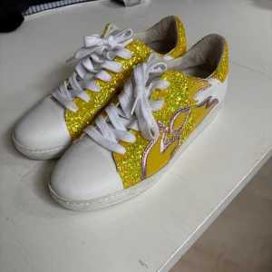 Sneakers från Custommade - Säljer ett par unika sneakers från Custommade med vit bas och stora partier i gul glitter. Det är endast använda fåtal gånger. Skorna har vita snören och en cool lila och rosa flammdetalj på sidorna. Perfekt för studenten eller liknande. Nypris ligger på ca 2500. 
