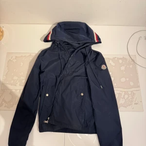 Mörkblå vindjacka från Moncler - Snygg mörkblå vindjacka från Moncler med huva och dragkedja. Jackan har vita och röda detaljer på huvan, två stora fickor framtill med knappar och Moncler-logga på ärmen. Perfekt för blåsiga dagar och enkel att matcha.