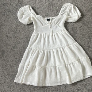 Vit volangklänning med puffärm från Gina Tricot - Superfin vit klänning från Gina Tricot med puffärmar och volanger. Klänningen har v-ringning och är sydd i ett lätt, strukturerat tyg. Perfekt för sommarens alla tillfällen!