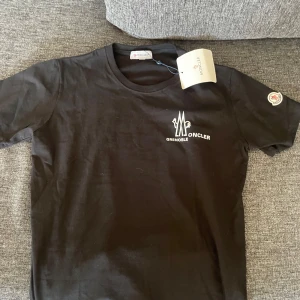 Svart t-shirt från Moncler Grenoble - Säljer en svart t-shirt från Moncler Grenoble med vit logga tryckt på bröstet och Moncler-märke på ärmen. Klassisk rund hals och korta ärmar. Perfekt för en stilren och enkel look. Storlek M men passar S lika fint