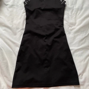 Svart kort klänning med nitar och detaljer - Säljer en svart kort klänning med unika banddetaljer och silvriga nitar längs axelband och rygg. Klänningen har en tight passform och är ärmlös med coola cut-outs upptill. Vet inte exakta storleken men passar xs-d