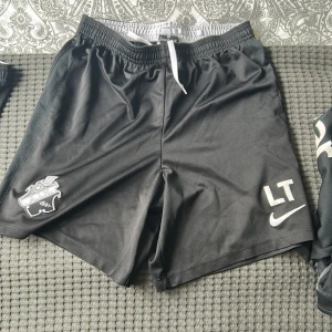 Svarta fotbollsshorts från Nike med AIK-logga - Svarta fotbollsshorts från Nike med AIK:s klubbmärke på vänster ben och initialerna LT samt Nike-logga på höger ben. Resår och vit dragsko i midjan för justerbar passform. Perfekta för fotbollsträning eller match.