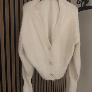 Vit kofta med glittriga knappar - Säljer en fluffig och mjuk vit kofta med djup v-ringning och tre glittriga dekorativa knappar framtill. Koftan har långa ärmar.