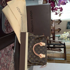 Louis Vuitton Monogram handväska - Säljer en klassisk handväska från Louis Vuitton i brunt monogrammönster med ljusa handtag och guldfärgade metalldetaljer. Väskan har dragkedja och är tillverkad i textil med detaljer i vachettaläder. Kommer med dustbag och originalkartong.