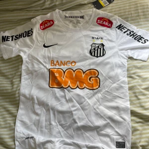 Vit Santos fotbollströja Neymar Jr #11 - Santos FC matchtröja i vitt med Neymar Jr och nummer 11 på ryggen. Tröjan har korta ärmar, svarta och orangea tryck, samt flera sponsorer. Nike-logga och Dri-FIT material för bra andningsförmåga. Tröjan är från säsong 2011 storlek s men passar m