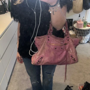 Rosa Balenciaga City Bag - Cool rosa handväska från Balenciaga med klassiska nitar och dragkedjedetaljer. Väskan har ett stort huvudfack, ytterficka med dragkedja och dekorativa remmar på handtagen. Perfekt för dig som gillar statement-accessoarer. Väskan är använd mycket och därav effekterna på handtagen, men inget som stör när man använder den!💕