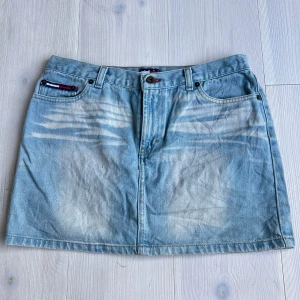 Ljusblå jeanskjol med broderade bakfickor - Säljer en ljusblå jeanskjol från Tommy Hilfiger med klassisk femficksdesign och röda broderier på bakfickorna. Kjolen har bälteshällor och knappgylf framtill. Perfekt till sommaren! Midjemåttet är 39cm rakt över och längden är 34cm