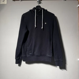 Morris hoodie - Säljer en svart hoodie med vit dragsko och liten broderad detalj på bröstet. Tröjan har huva och långa ärmar, samt en klassisk känguruficka framtill. Perfekt för en avslappnad stil. Tröjan är från Morris. Skriv om du har några funderingar.