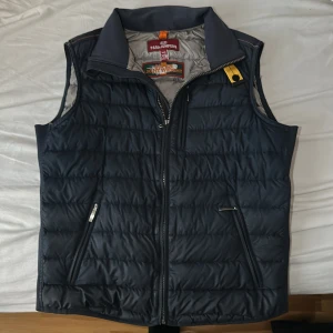 Mörkblå dunväst från Parajumpers - Säljer en mörkblå dunväst från Parajumpers med två dragkedjefickor framtill och klassisk gul detalj vid kragen. Västen är superlätt och har quiltad design samt hög krage. Perfekt för lager-på-lager under kyliga dagar.