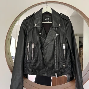 Svart skinnjacka från Zara - Säljer en svart skinnjacka från Zara med klassisk bikerstil. Jackan har silvriga dragkedjor, axelklaffar och tryckknappar. 100% läder, Originalpris 1399kr