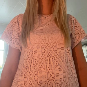 Ljusrosa spetstopp med korta ärmar - Säljer en ljusrosa topp med korta ärmar och snyggt spetsmönster över hela plagget. Toppen har en rak passform och är tillverkad i ett mjukt bomullsmaterial. Perfekt för dig som gillar romantiska detaljer och vill ha en feminin look.