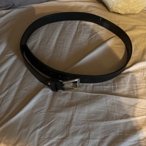 Svart klassiskt skärp i skinn - Snyggt svart bälte i skinn med stilren silverfärgad metallspänne. Perfekt för att ge en clean och tidlös look till dina outfits. Bältet har flera hål för justerbar passform och passar både jeans och kostymbyxor.