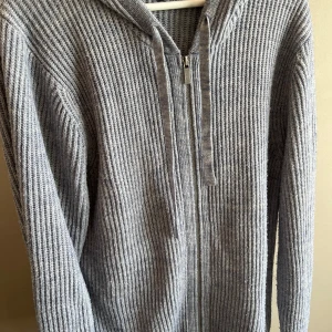 Grå hoodie från Lawrence Grey - Säljer en grå ribbad hoodie med huva och snörning. Tröjan har dragkedja framtill och är stickad i ett mjukt material som känns skönt mot huden. Långärmad och perfekt för kyliga dagar. Enkel och stilren design som funkar till många olika outfits.