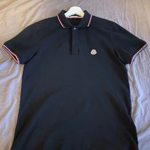 Svart Moncler pikétröja med röd/vit kant - Svart pikétröja från Moncler med klassisk krage och korta ärmar. Tröjan har snygga rödvita ränder på krage och ärmslut samt Moncler-logga på bröstet. Tillverkad i bomull och har knappar framtill. Perfekt för en clean och sportig look.