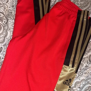Röda Adidas mjukisbyxor med guld - Säljer ett par röda Adidas mjukisbyxor med svarta och guldiga detaljer längs sidorna. Byxorna har elastisk midja och klassiska Adidas-ränder i guld. Materialet är mjukt och skönt, perfekt för chill eller träning. Adidas-logga i svart på benet. Strl S herr