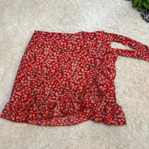 Röd blommig omlottkjol med volang -  röd omlottkjol med små vita och gula blommor. Kjolen har volang längst ner och knyts i midjan 