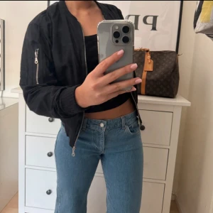 Svart bomberjacka med dragkedja - Säljer en svart bomberjacka med coola detaljer som dragkedja på ärmen och ribbade muddar. Jackan har klassisk rund hals och är helsvart, 