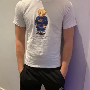 Vit Polo Bear t-shirt från Ralph Lauren - Vit t-shirt från Ralph Lauren med ikonisk Polo Bear-tryck på framsidan. Björnen har en blå tröja med amerikansk flagga och jeans. T-shirten har korta ärmar, rund hals och Polo-logga på ryggen. Skön bomullskvalitet och normal passform. Modellen är 180cm och väger 73kg neck tagen är avklippt och därav det billiga priset 