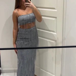 Silver glittrig topp o kjol med fransar - Säljer en silverfärgad set med tunna axelband och glittriga paljetter. Settet har fransdetaljer som ger extra rörelse och en öppen midja som visar lite hud. Perfekt för dig som vill sticka ut på festen!