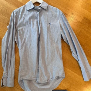 Blårandig Burberry skjorta - Snygg blårandig skjorta från Burberry Brit med klassisk krage och långa ärmar. Insidan av kragen och manschetterna har det ikoniska Burberry-rutiga mönstret i beige, svart och rött. Skjortan har knappar och diskret logobroderi på bröstet. Perfekt för dig som gillar stilrena detaljer.