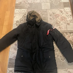 Svart parkajacka Alpha Industries M - Parkajacka från Alpha Industries i svart med huva och mjukt brunt foder. Jackan har flera fickor, dragkedja och en ikonisk röd 'Remove Before Flight'-tag på ärmen. Snyggt broderad patch på axeln och tryck på ryggen. Perfekt för kalla dagar.