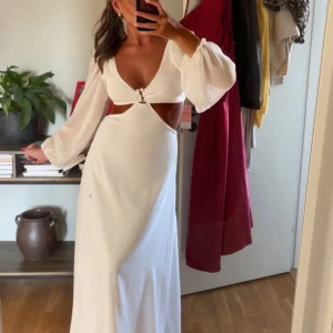 Vit långklänning - Säljer en vit långklänning med ballongärmar och snygga cut-outs vid midjan. Klänningen har djup v-ringning och ringdetalj framtill. Materialet är lätt och faller fint, perfekt för dig som vill ha en trendig och elegant look.