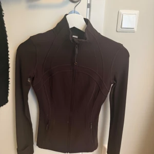 Brun träningsjacka från Lululemon och träningstights - Säljer en brun, figursydd träningsjacka från Lululemon med hel dragkedja framtill och höga sömdetaljer. Jackan har lång ärm, ståkrage och diskret logga på ryggen och ärmen. Perfekt för dig som vill ha en sportig och snygg outfit, har endast använt detta sett 2 gånger de sitter jätte fint på, säljer på grund av ingen användning (ni kan köpa ett plagg separat) 