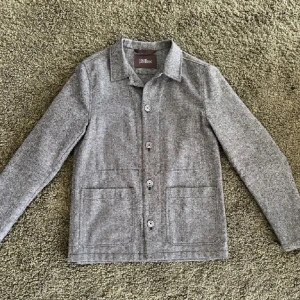 Grå overshirt från Oscar Jacobson - Snygg grå overshirt från Oscar Jacobson med klassisk krage och stora framfickor. Jackan har knappar hela vägen och är tillverkad i mjuk ullblandning. Perfekt för lager-på-lager och passar dig som gillar stilren och enkel design.