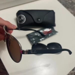 Ray-Ban solglasögon  - Ray-Ban solglasögon med silverfärgad metallram och svarta skalmar. Köpta från synoptik för många år sen, men knappt använda. Köpta med min styrka på glaset, men fick med ett par extra linser utan styrka så man kan använda dom som vanliga solglasögon. Dom extra glasen är mörkgråa. Det vänstra glaset fick en liten repa på insidan, då jag råkade tappa den när jag skulle fota (syns lite på fjärde bilden, nere vid vänster kant). Kommer med original fodral.