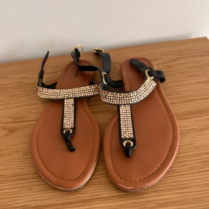 Svarta sandaler med strass från Graceland - Snygga svarta sandaler från Graceland med bruna sulor och glittriga strassdetaljer på remmarna. Sandalerna har guldfärgade spännen och platt sula med dekorativt mönster undertill. Perfekta för att addera lite bling till din sommarstil!