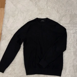 Svart stickad tröja från J.Lindeberg - Svart stickad tröja från J.Lindeberg i storlek S. Klassisk rund hals och långa ärmar, perfekt för dig som gillar stilrena och enkla plagg. Materialet är mjukt och skönt, passar bra till jeans eller chinos. Snyggt ribbad vid hals, ärmslut och nederkant.