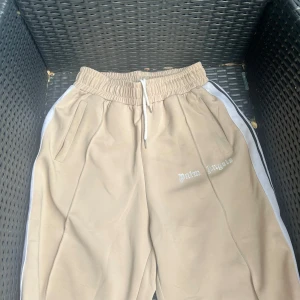 Beige Palm Angels mjukisbyxor med ränder - Snygga beige mjukisbyxor från Palm Angels med vita och svarta ränder längs sidorna. Byxorna har elastisk midja med vit snörning och sidofickor. Palm Angels-loggan är broderad på framsidan. Perfekta för chill eller streetwear.