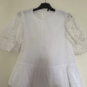 Vit blus med spetsärmar från Zara - Supersöt vit blus från Zara med puffiga spetsärmar och rund halsringning. Blusen har en volang nedtill som ger en extra touch och är tillverkad i mjuk bomull. Använd endast 1 gång. 