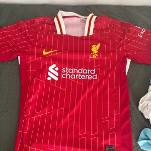 Liverpool FC röd Nike matchtröja - Liverpool FCs officiella matchtröja från Nike i röd polyester med gula detaljer och tunna gula ränder. Kortärmad modell med vit och gul krage, broderat klubbmärke och tryckt sponsorlogga på bröstet. Snygg design för dig som älskar fotboll och LFC. THAI KVALITÉ!!!