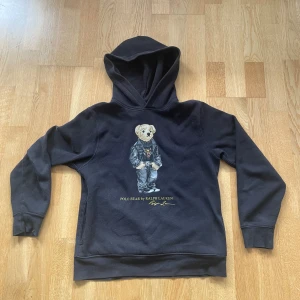 Svart Polo Bear hoodie från Ralph Lauren - Svart hoodie från Polo Ralph Lauren med ikoniska Polo Bear-trycket på bröstet. Tröjan har huva, långa ärmar och ribbade muddar. Materialet är mjuk bomull och polyesterblandning, perfekt för chill dagar. Snygg och klassisk streetwear-vibe med en cool björn i kostym framtill. Säljer pga att den blivit för liten men så länge ja användt den så har den varit hur skön och go som helst rikigt snygg också. Väldigt bra skick och inga konstigheter me den. Hör av er vid minsta fundering kan diskutera pris😊