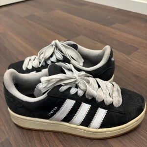 Adidas Campus svarta sneakers - Adidas Campus sneakers i svart mocka med klassiska vita ränder på sidorna och vit snörning. Låg modell med rund tå och platt sula. Snygg retrostil som funkar till jeans eller shorts. Campus-logga på sidan och mjuk innersula för extra komfort.